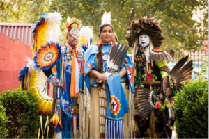 Three people stand outdoors dressed in distinctive, colorful, indigenous outfits.