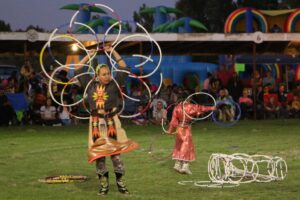 A hoop dance artist and her daughter dressed in traditional clothes perform for an audience at an outdoor venue.