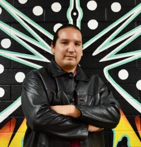 Artist Michael Elizondo, Jr. standing with a smile and folded arms in front of a colorful artwork painted on a brick wall.