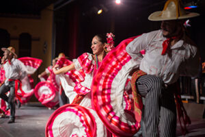 Several traditional Mexican dancers twirl their voluminous white-and-red skirts while two male dancers are shown raising their right knees with their hands behind their backs.