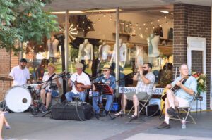 Six street musicians play instruments and sing while seated by a brightly lit storefront. 